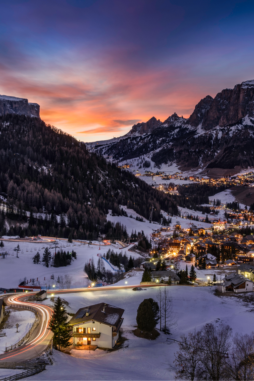 Evening view over a snow-covered alpine village and winding mountain roads in the French Alps, with illuminated houses beneath rugged peaks